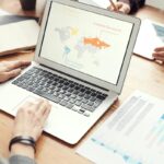 Laptop showing a slide with a world map and three labeled points, with printed charts, notebook and tablet on a desk