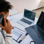 Salesperson on the phone reviewing printed charts and two laptops before a sales meeting
